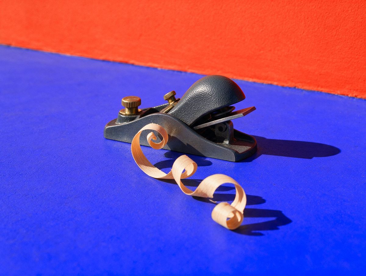 A block plane beside one long curl of pale wood shaving on a cobalt floor against a vermilion wall: the tools do the heavy lifting, the finish is done by hand.
